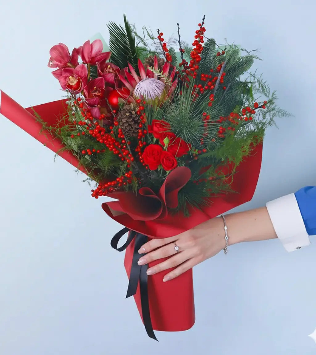 Crimson Celebration luxury Christmas bouquet with pink cymbidium orchids, red roses, protea blooms, and festive greenery wrapped in red and black paper Dubai