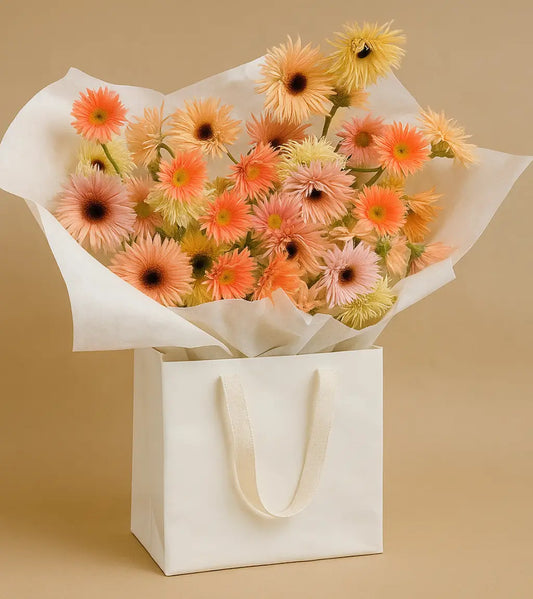 Soft pastel Gerbera daisies in peach, yellow, and cream arranged in a light brown kraft bag with black ribbon handles – Pastel Breeze flower arrangement, Dubai.